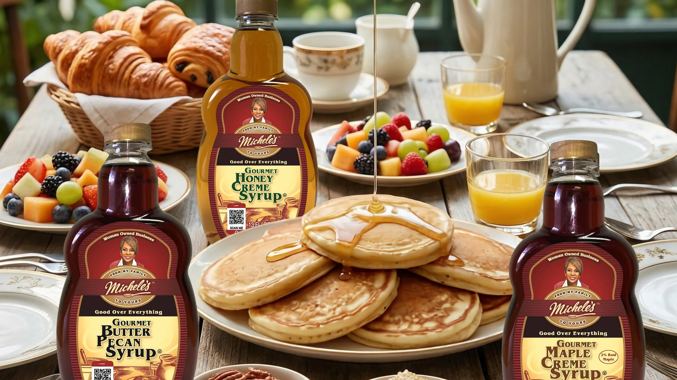 Plate of pancakes with syrup pouring from above, surrounded by Michele’s honey, maple, and butter pecan syrups, fruit, croissants, and orange juice.